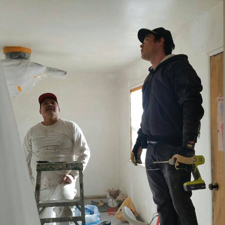 Two construction workers inspect an unfinished interior room with white drywall and plastic sheeting