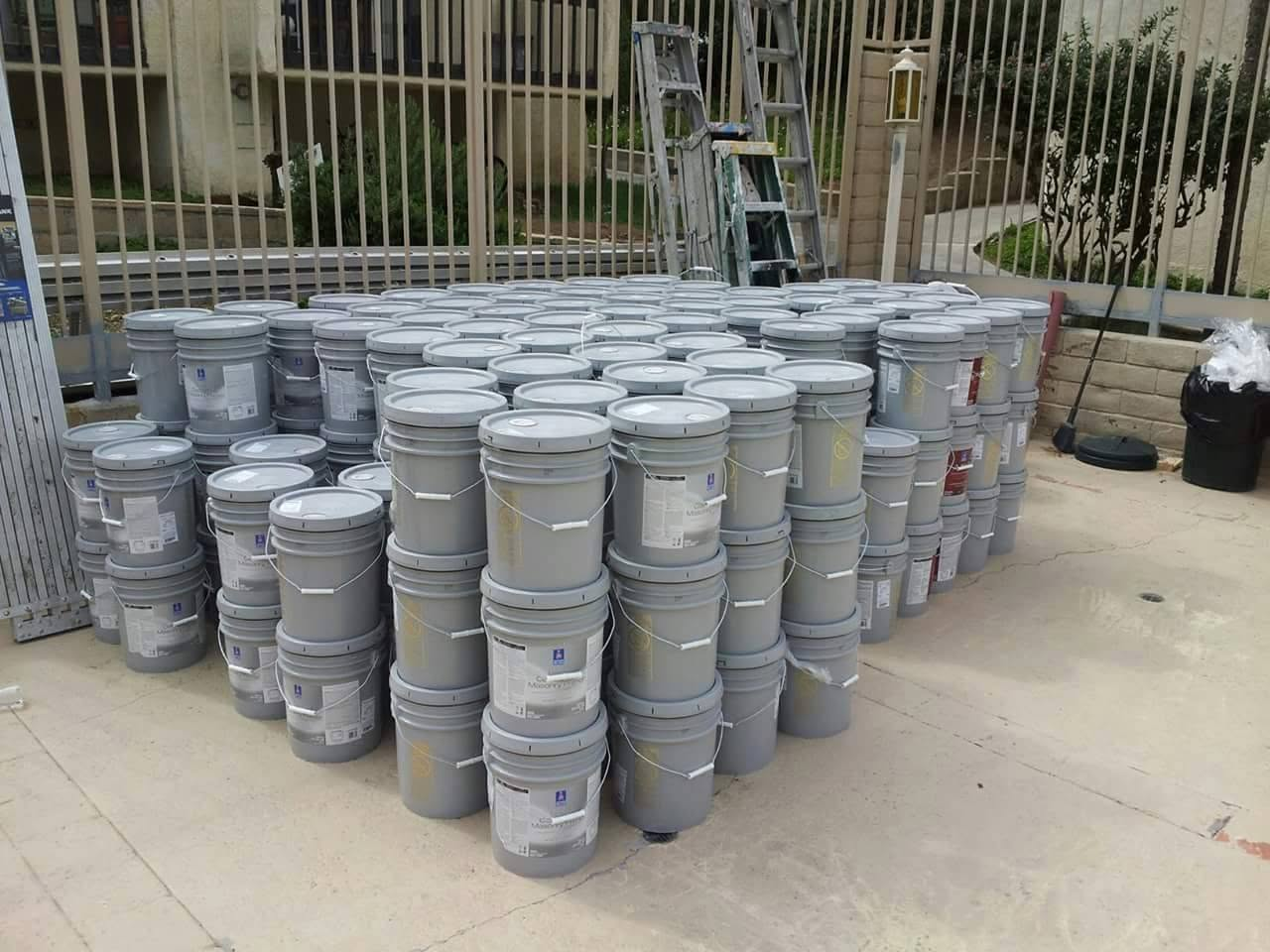 Large collection of gray industrial paint buckets stacked in a courtyard with metal gates and ladder visible