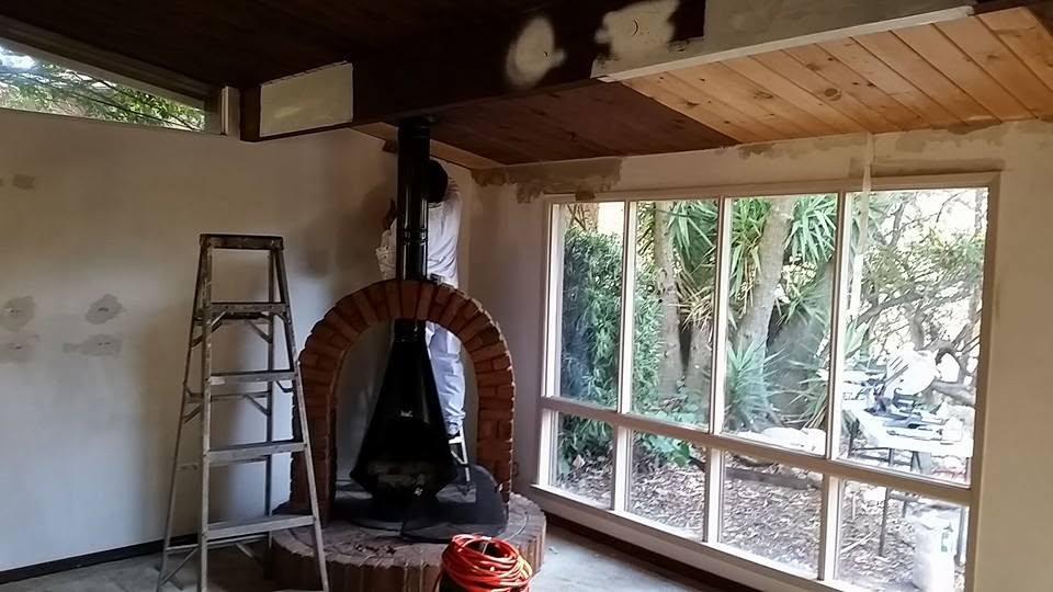 Interior construction space with brick fireplace, ladder, and large windows overlooking tropical garden